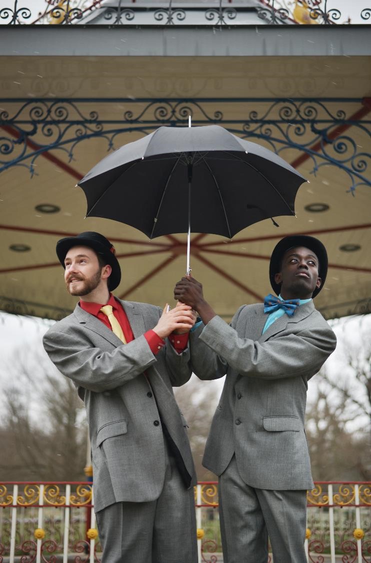 Two male dancers in suits holding an umbrella above their heads